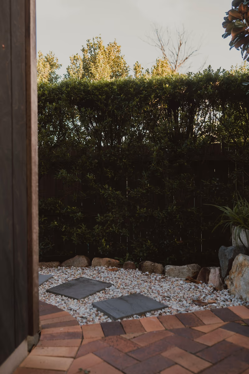 Garden corner with a herringbone brick paver terrace, stepping stones set into a gravel bed, boulder edging and a clipped screening hedge along the fence