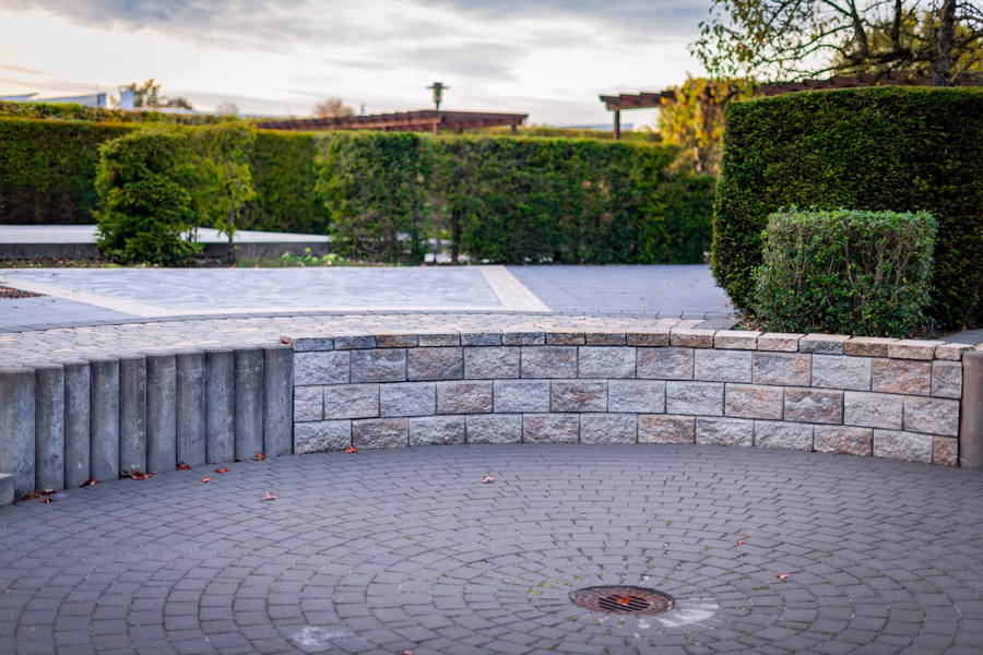 Low segmental block seat wall curving around a circular paver terrace, with a centre drain grate set into the paving and clipped hedges behind